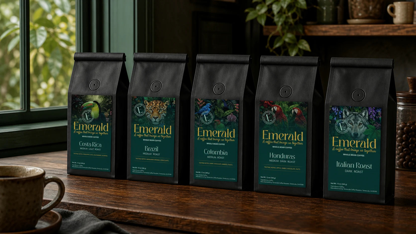 Five Emerald Cafe coffee bags lined up on a wooden counter — Costa Rica, Brazil, Colombia, Honduras and Italian Roast — beside a window with green foliage outside